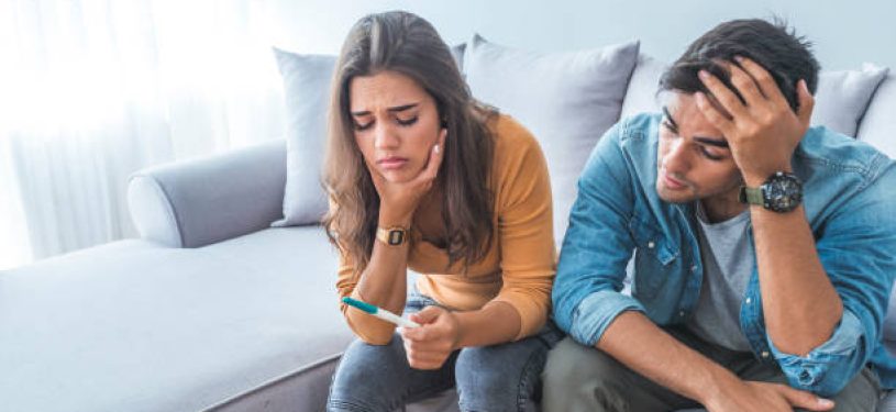 Sad stressed couple with pregnancy test sitting at home. Upset woman is looking in pregnancy test. Frustrated man is sitting next to her. Worried about the serious consequences
