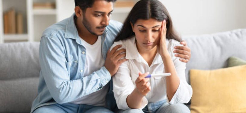 Unwanted pregnancy. Worried hindu couple looking at positive test result, stressed indian man and woman not ready to be parents, sitting on sofa at home