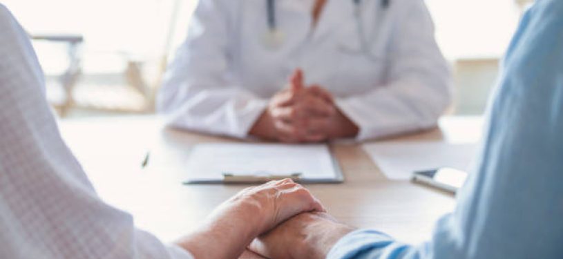 Mature couple holding hands at a doctors office. Doctor can be seen in the background.