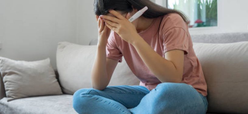 Sad woman sitting on sofa and holding pregnancy test with negative result